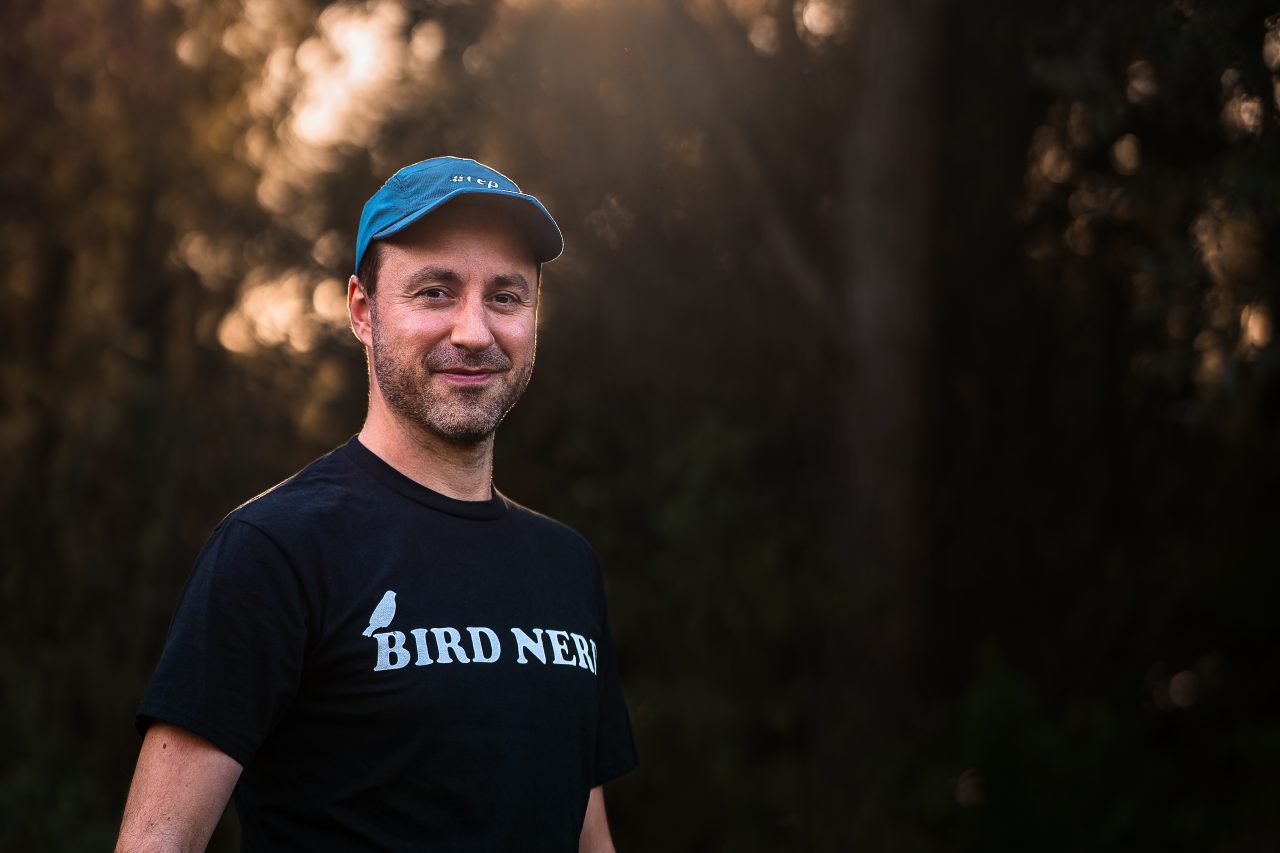 Jan Wittmer im Gegenlicht vor Bäumen, mit Basecap und einem T-Shirt mit der Aufschrift Bird Nerd